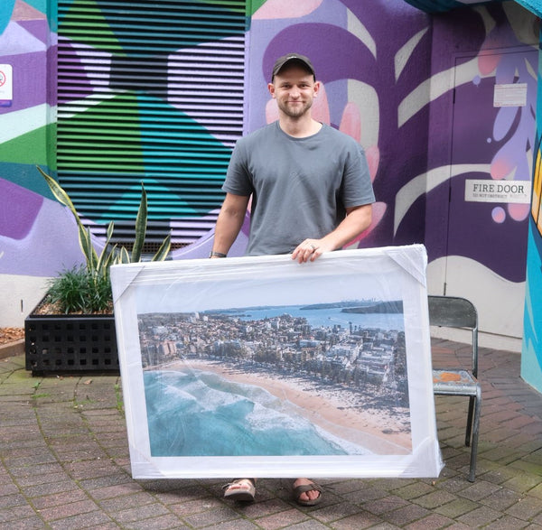 Guy holding Large Framed Artwork of Manly Beach in front of colorful purple graffiti wall