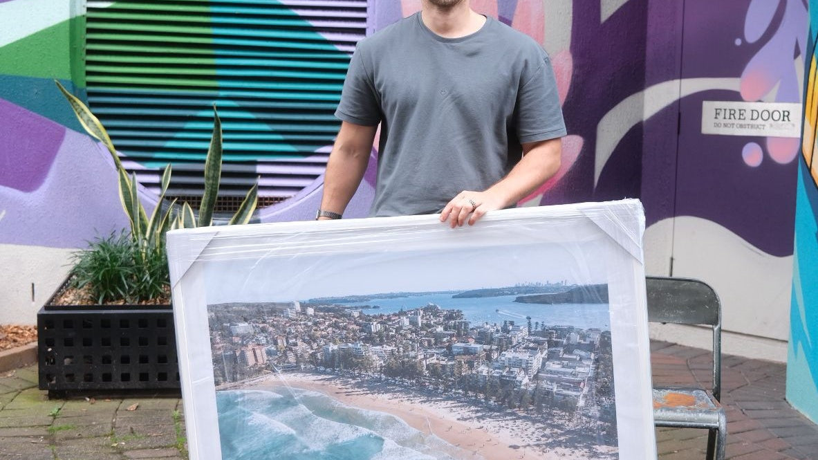 Guy holding Large Framed Artwork of Manly Beach in front of colorful purple graffiti wall
